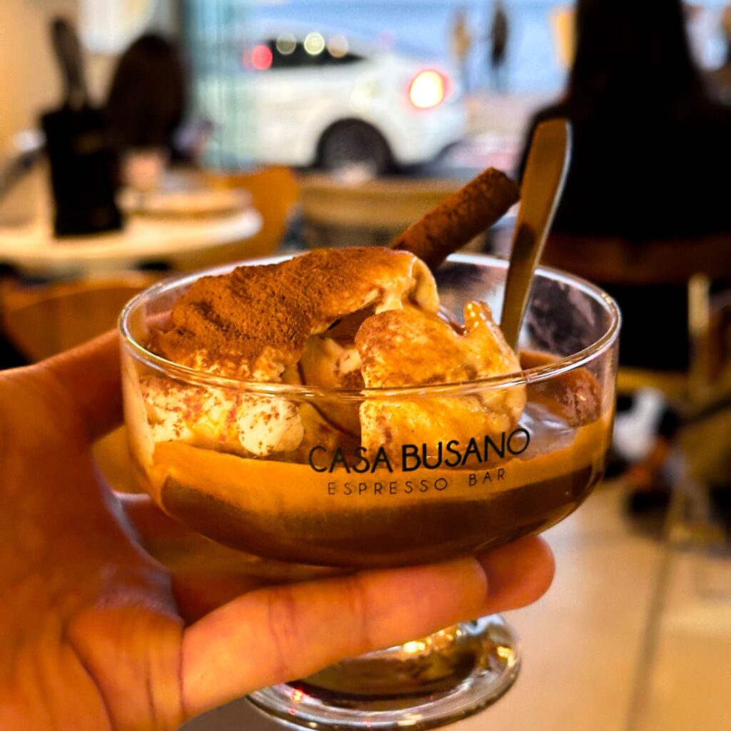 Hand holding a footed glass of cocoa-dusted cream dessert with a wafer roll and a spoon, printed with the CASA BUSANO logo