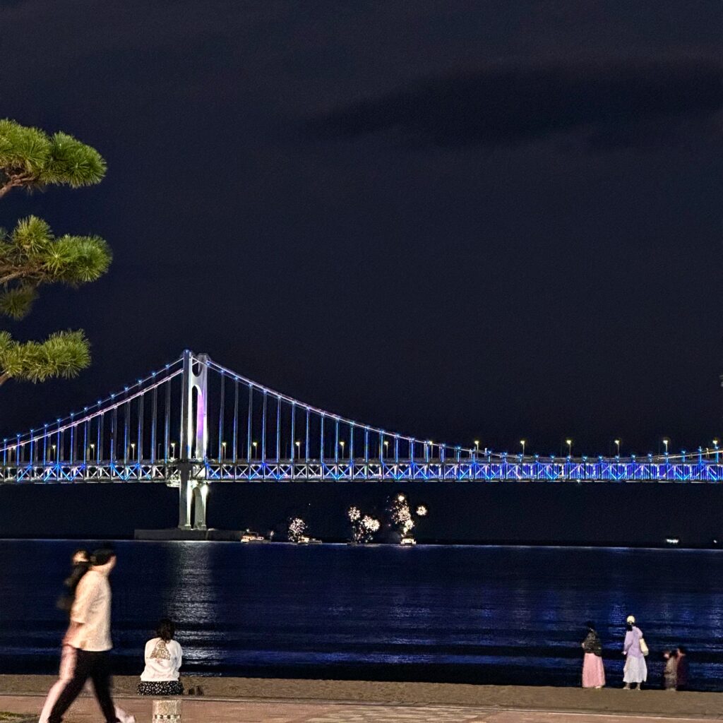 Gwangan Bridge lit in blue over the dark sea at night, with people walking along the beach