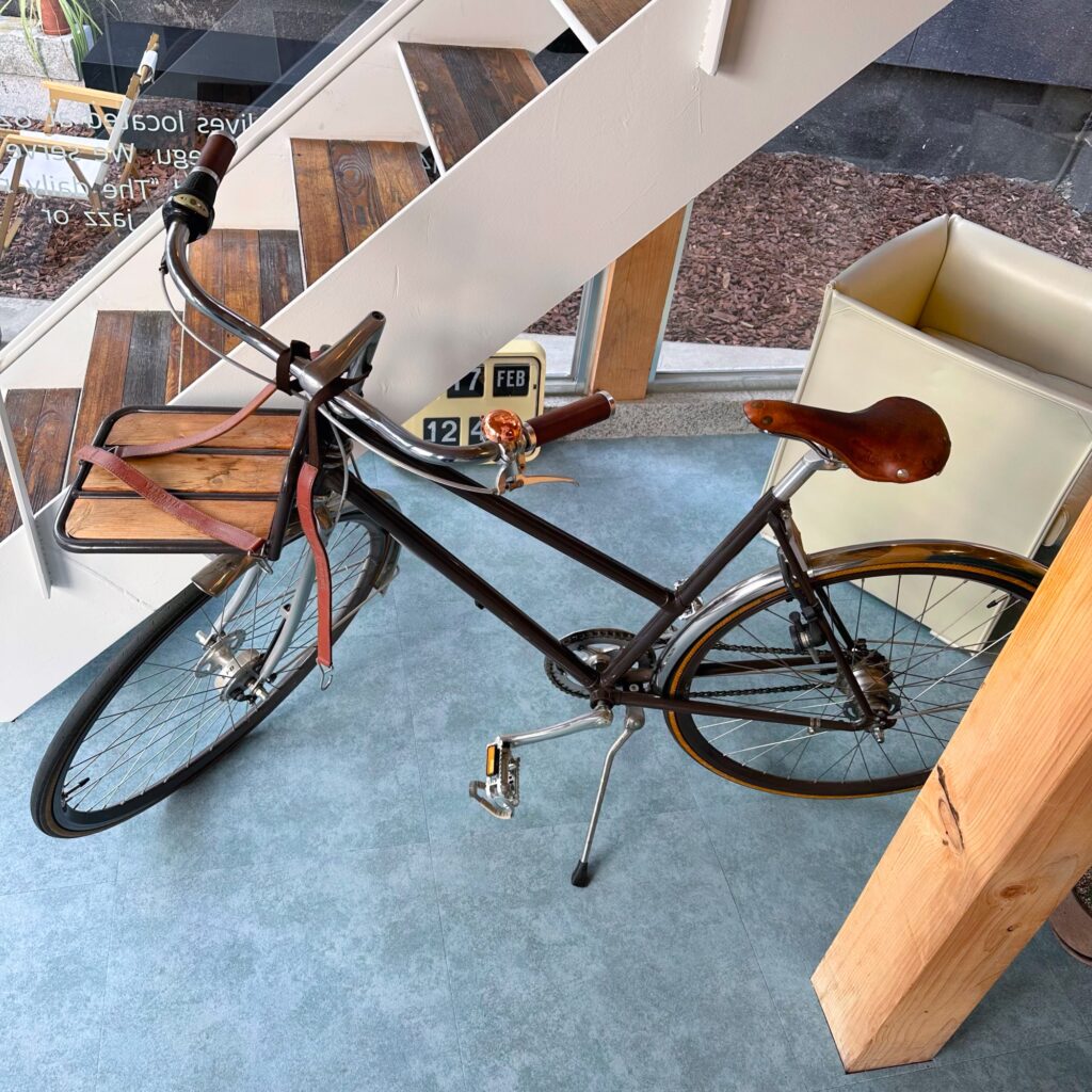 A vintage bicycle with a brown leather saddle and a wooden crate, parked under the white staircase