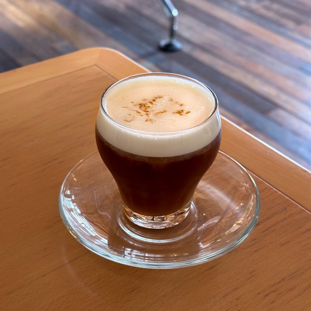 A coffee with a thick layer of foam and a light dusting on top, in a small glass on a glass saucer