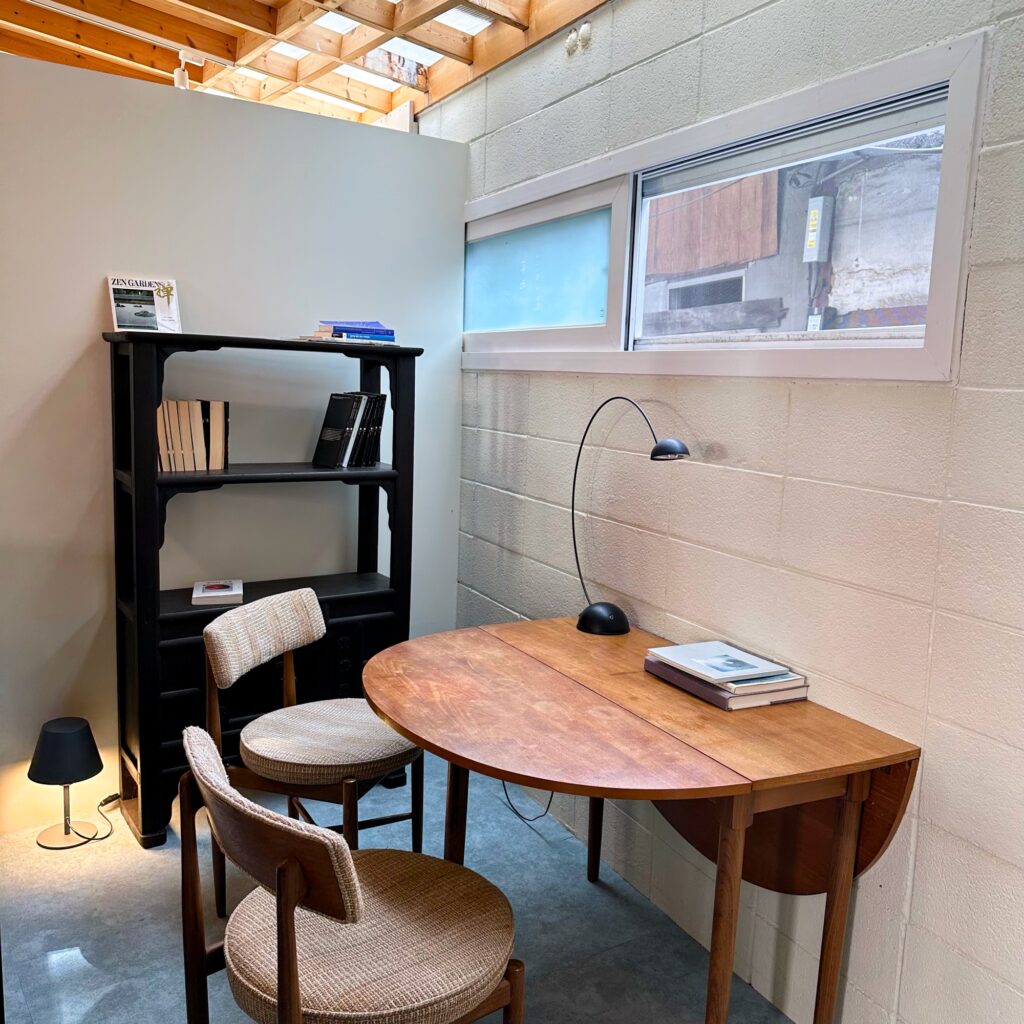 A quiet corner with a wooden drop-leaf table, a black desk lamp, two woven chairs and a black bookshelf