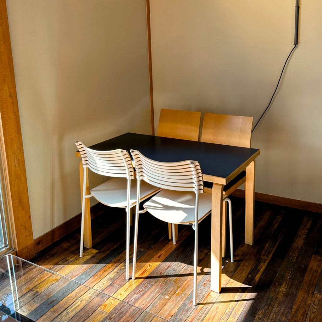 A black-topped table with white and light wood chairs in a sunny corner on a dark wooden floor
