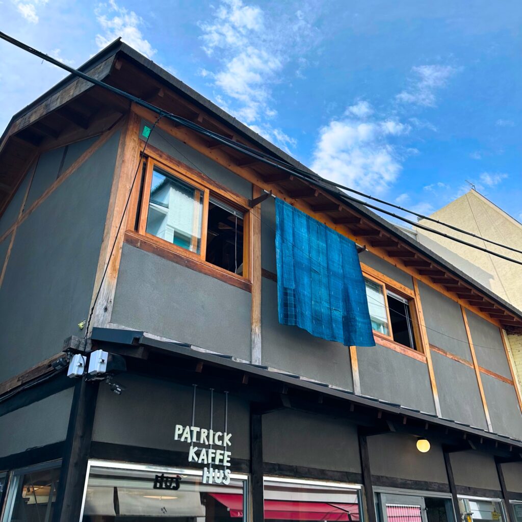 Two-story café with gray walls, wooden window frames and a blue cloth hanging from the upper window, with the Patrick Kaffe Hus sign under a blue sky